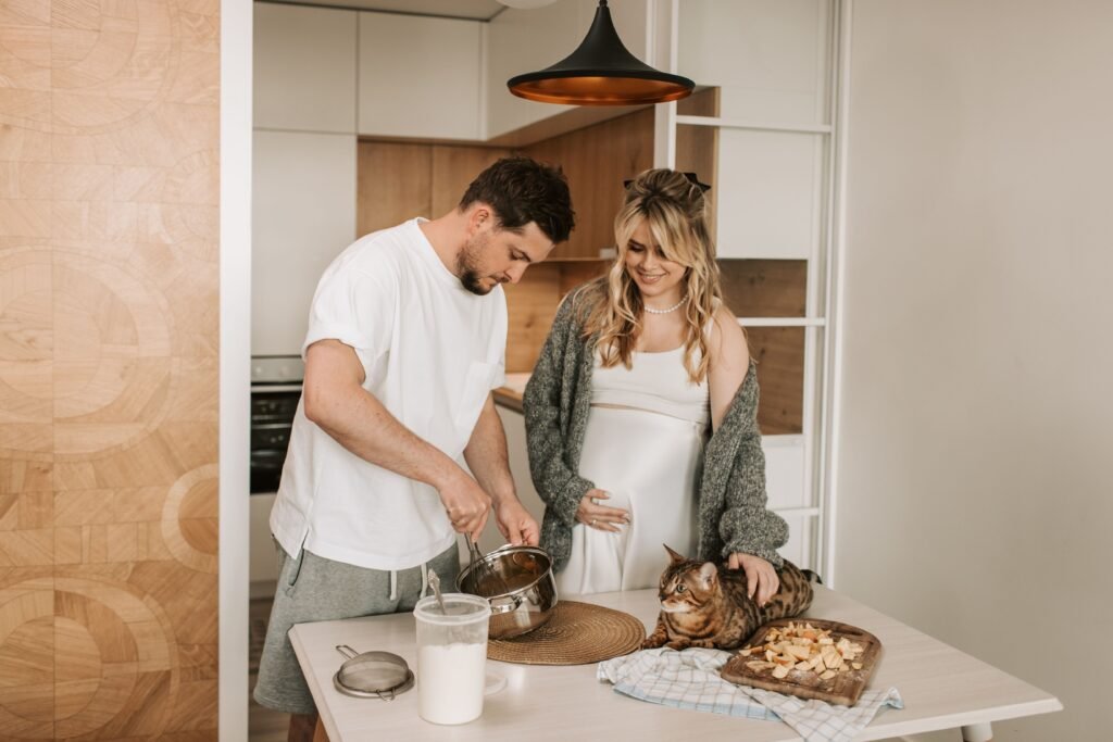 A pregnant couple bonding in a modern kitchen with their cat nearby and enjoying food preparation together.